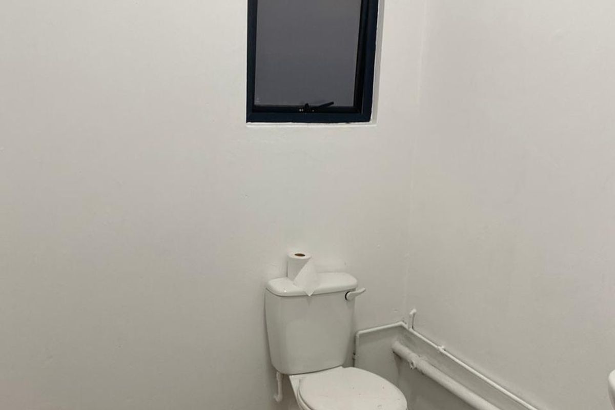 A functional industrial-style restroom featuring a standard toilet, exposed surface-mounted plumbing, and a small window for ventilation.