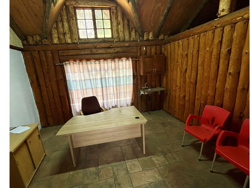 Rustic office space featuring log wall cladding, exposed timber rafters, tiled flooring, and a small wall-mounted sink with overhead cabinetry.