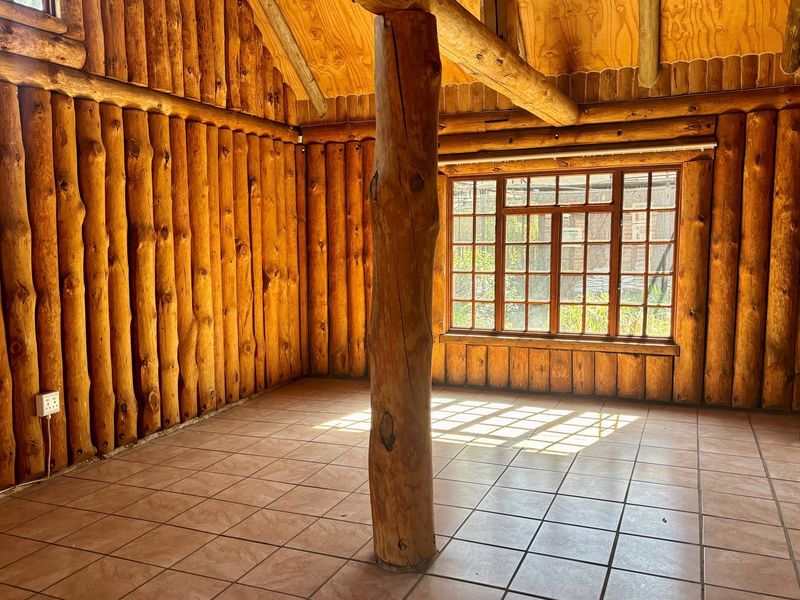Rustic interior space featuring exposed log walls, a central timber support pillar, tiled flooring, and large multi-pane windows providing natural light.