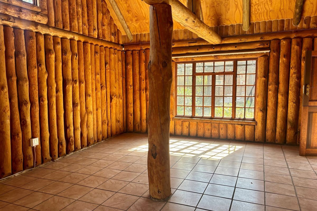 Rustic interior space featuring exposed log walls, a central timber support pillar, tiled flooring, and large multi-pane windows providing natural light.