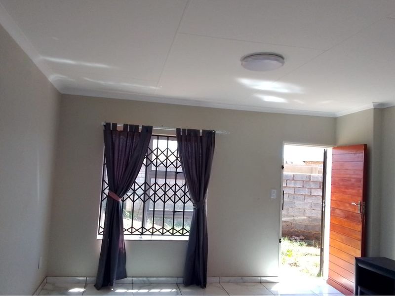 A bright, empty living room featuring tiled flooring, a security gate on the window, and direct access to the exterior through a wooden door.
