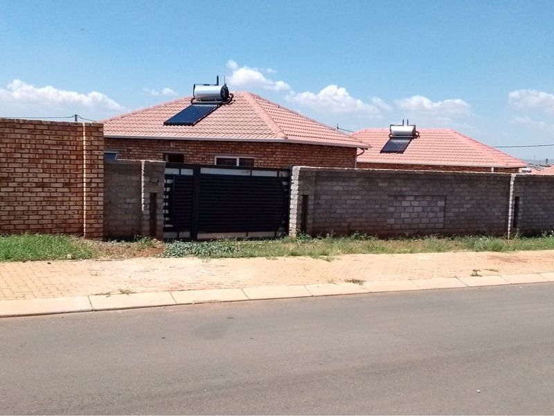 Front exterior view of a brick house featuring a tiled roof, solar water heating system, and a secure perimeter wall with a metal gate.