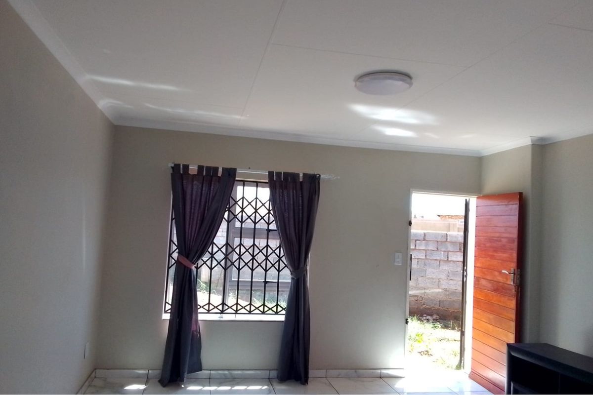 A bright, empty living room featuring tiled flooring, a security gate on the window, and direct access to the exterior through a wooden door.