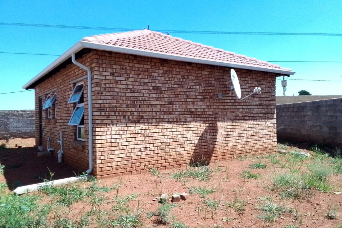 A side view of a face brick house featuring a tiled roof, aluminium windows, and a satellite dish, set on an unlandscaped plot.