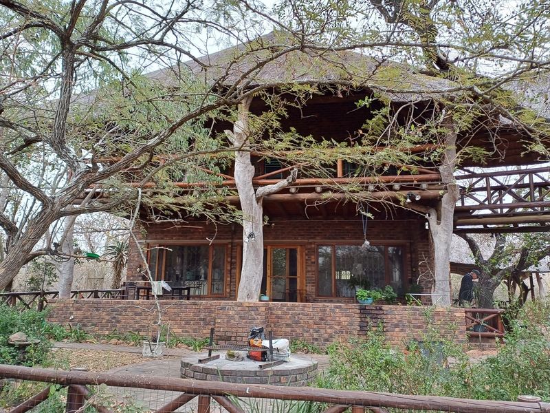 A rustic two-story house featuring a thatched roof, brick exterior, and a spacious timber balcony overlooking a garden with a built-in braai area.