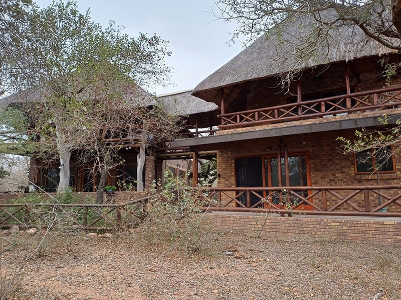 A two-story face brick house featuring a traditional thatched roof, timber balcony railings, and a covered patio area set in a natural bushveld environment.