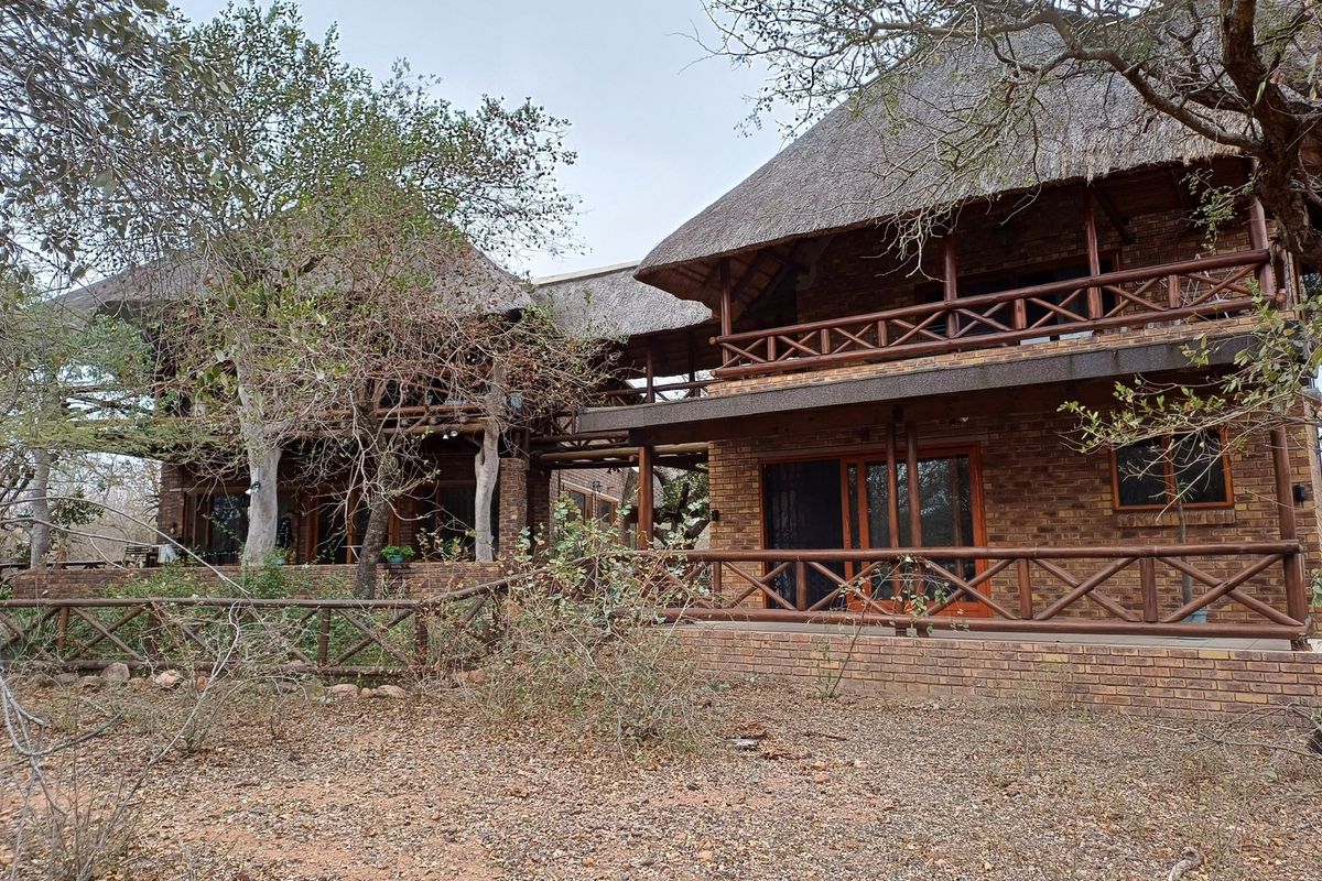 A two-story face brick house featuring a traditional thatched roof, timber balcony railings, and a covered patio area set in a natural bushveld environment.