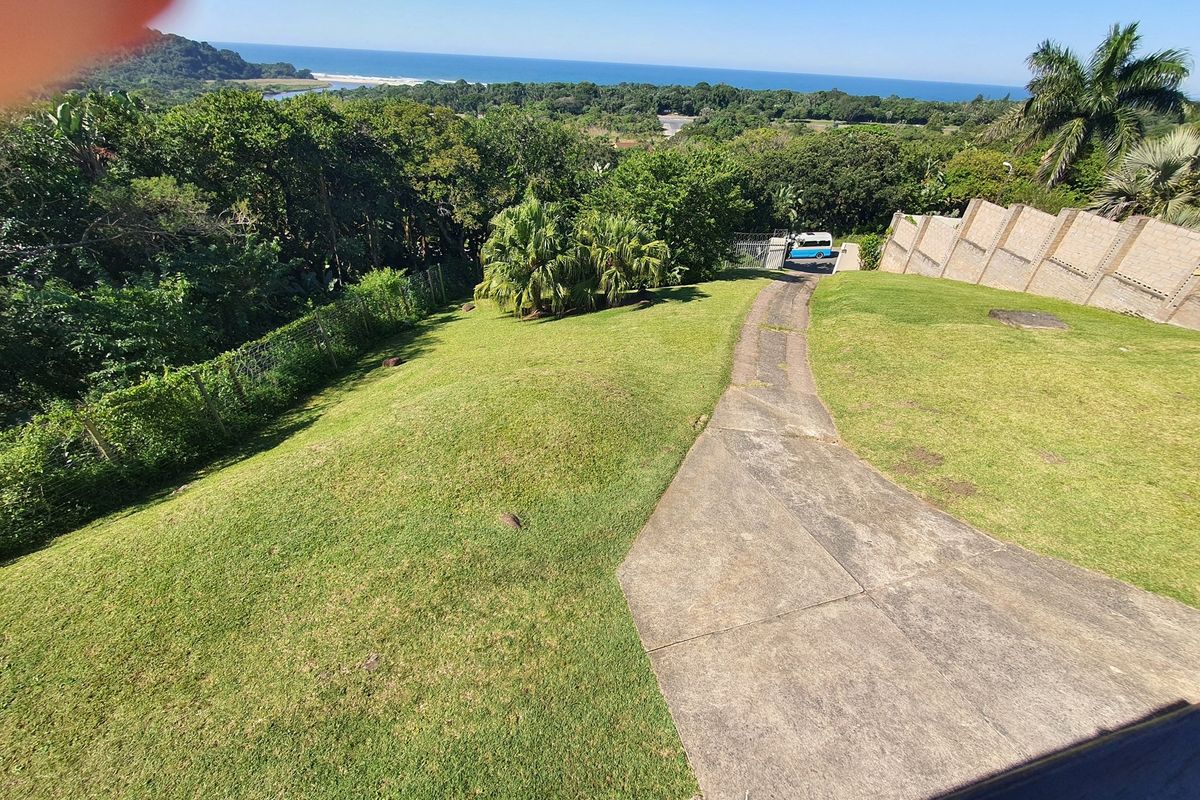 Elevated view of the property grounds featuring a sloping lawn, concrete driveway, and scenic coastal landscape in the background.