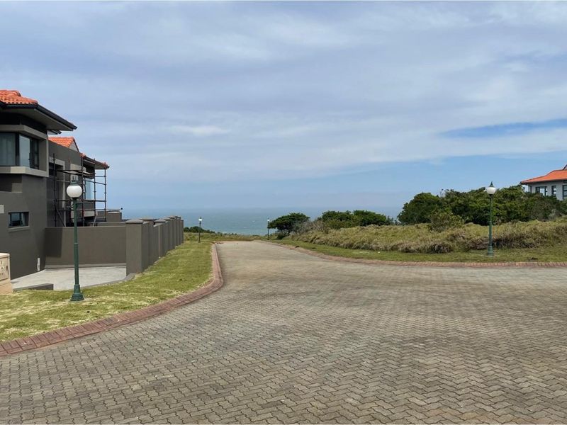 A view of a paved residential street leading towards the ocean, featuring landscaped verges, boundary walls, and nearby residential properties.