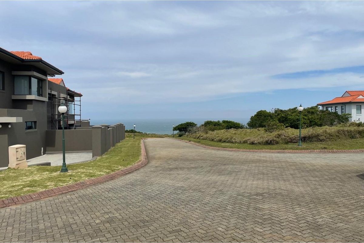 A view of a paved residential street leading towards the ocean, featuring landscaped verges, boundary walls, and nearby residential properties.