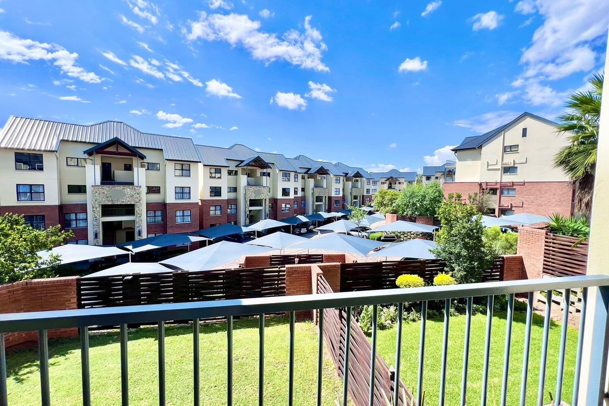 View from a balcony overlooking the apartment complex grounds, featuring landscaped gardens, paved walkways, and covered parking bays.