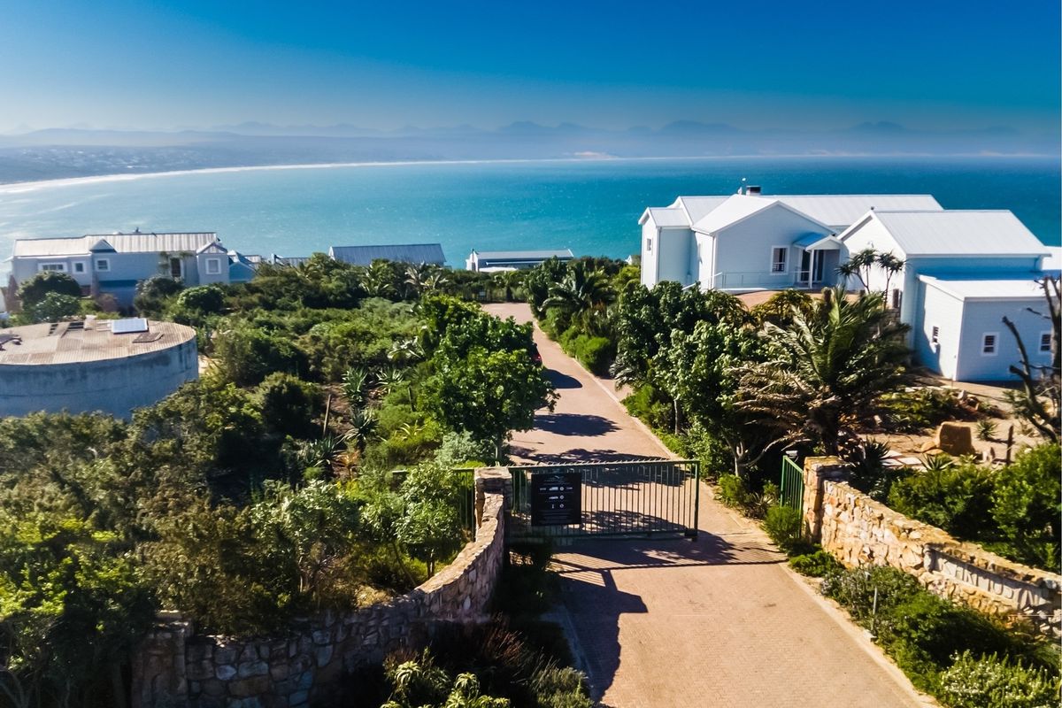 Aerial view of a residential property entrance featuring a paved driveway, stone boundary walls, and a metal security gate with coastal ocean views.