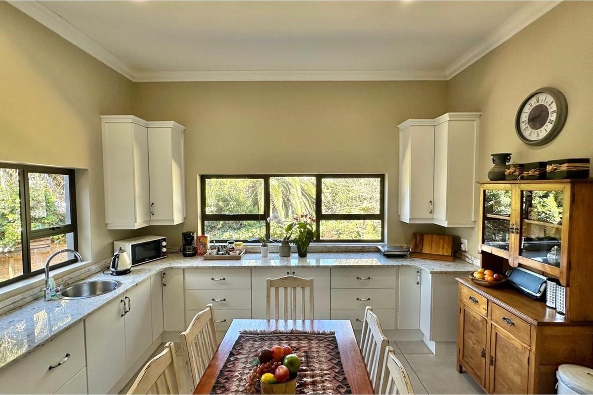 A bright kitchen featuring white cabinetry, granite countertops, and a stainless steel sink, with large windows providing natural light and garden views.