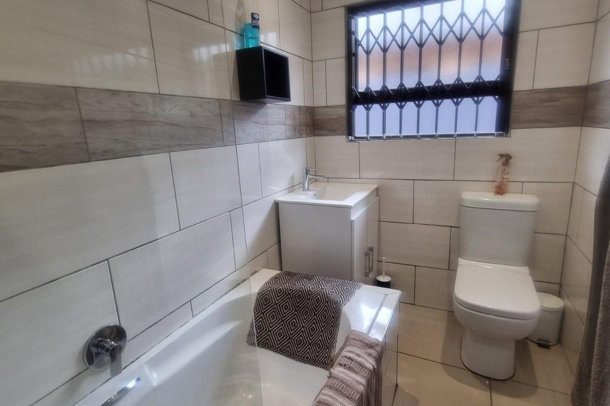 A modern bathroom featuring floor-to-ceiling wall tiles, a built-in bathtub with chrome fixtures, a vanity basin, and a toilet with a security-grilled window.