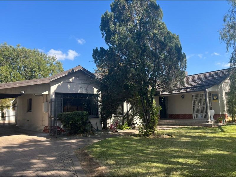 Front elevation of a single-story house featuring a paved driveway, attached carport, and established garden with mature trees.