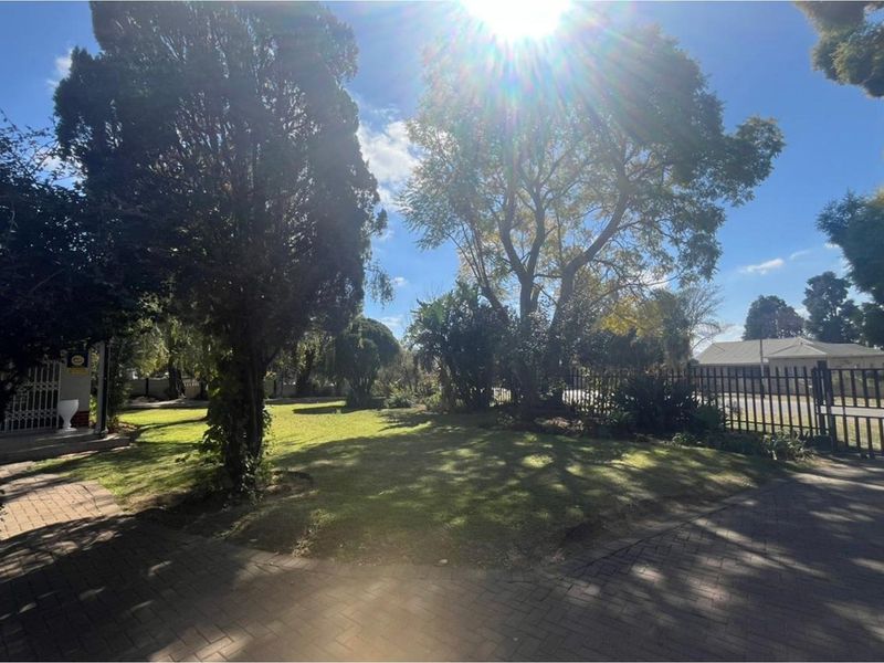 A well-maintained front garden featuring a paved driveway, established mature trees, and secure metal perimeter fencing.