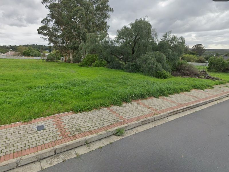 A vacant residential plot featuring established trees and a paved sidewalk along the street frontage.