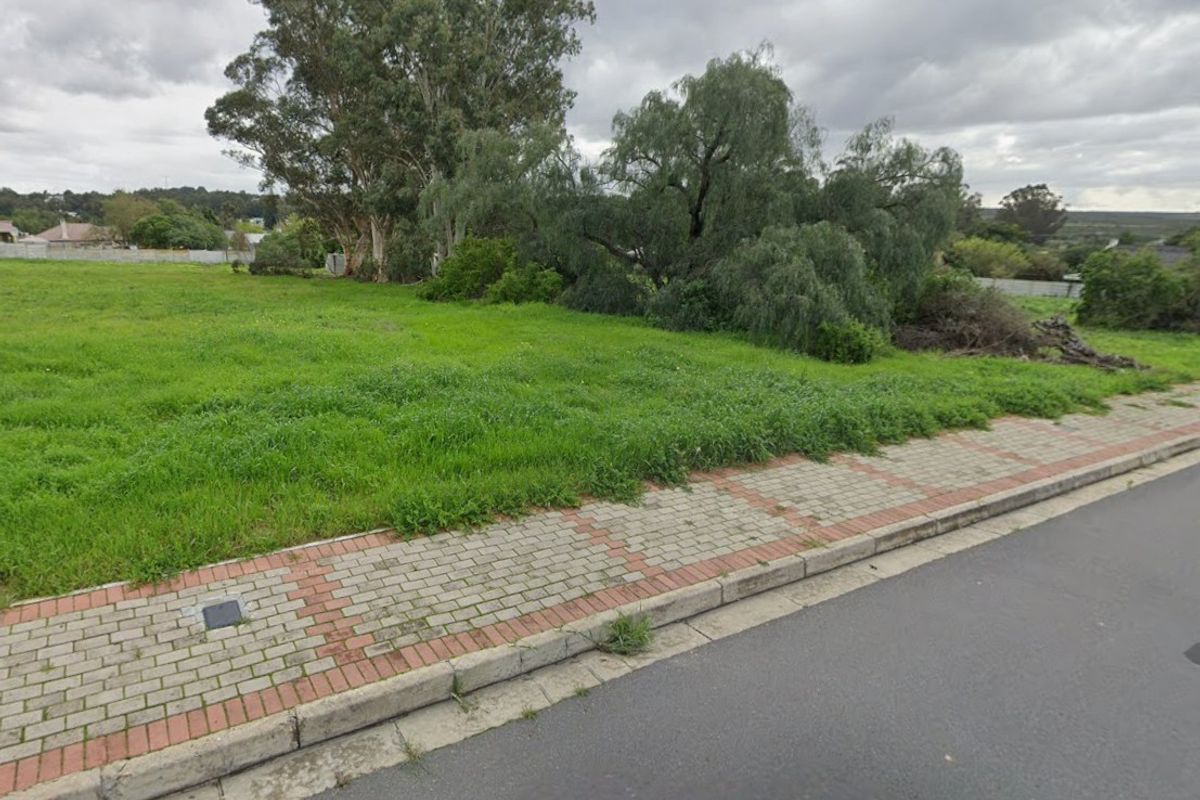 A vacant residential plot featuring established trees and a paved sidewalk along the street frontage.