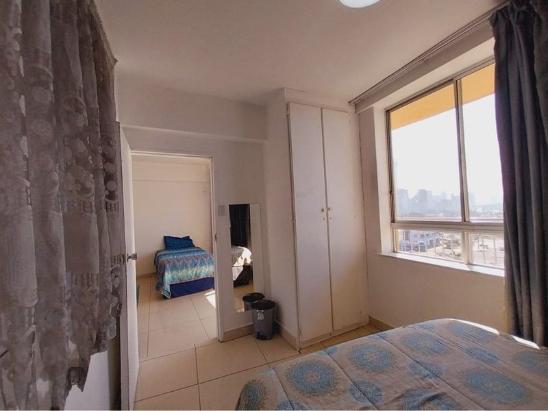 A bedroom featuring built-in cupboards, tiled flooring, and large windows providing natural light and urban views, with an adjacent room visible through the doorway.