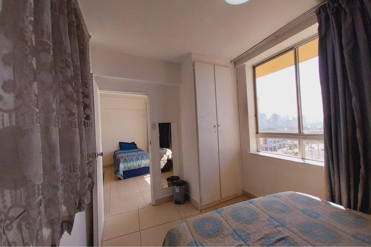 A bedroom featuring built-in cupboards, tiled flooring, and large windows providing natural light and urban views, with an adjacent room visible through the doorway.