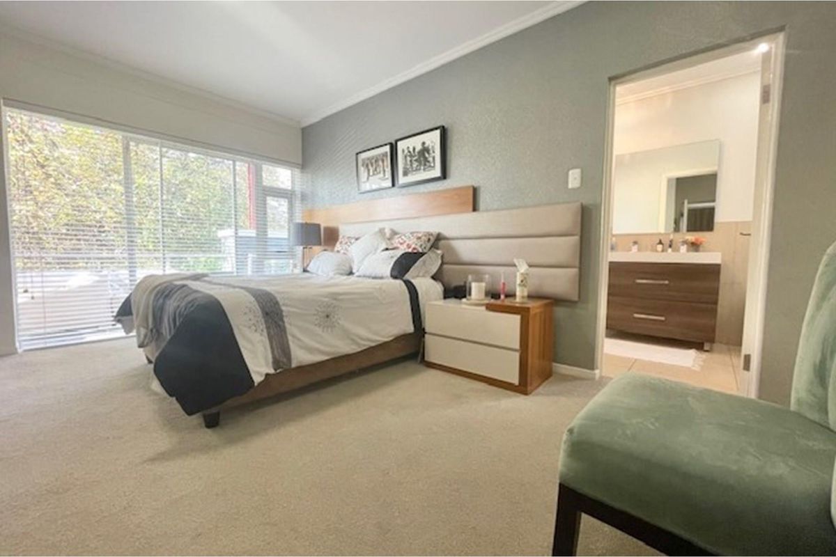A well-maintained bedroom featuring a built-in headboard, carpeted flooring, large windows with blinds, and an en-suite bathroom.