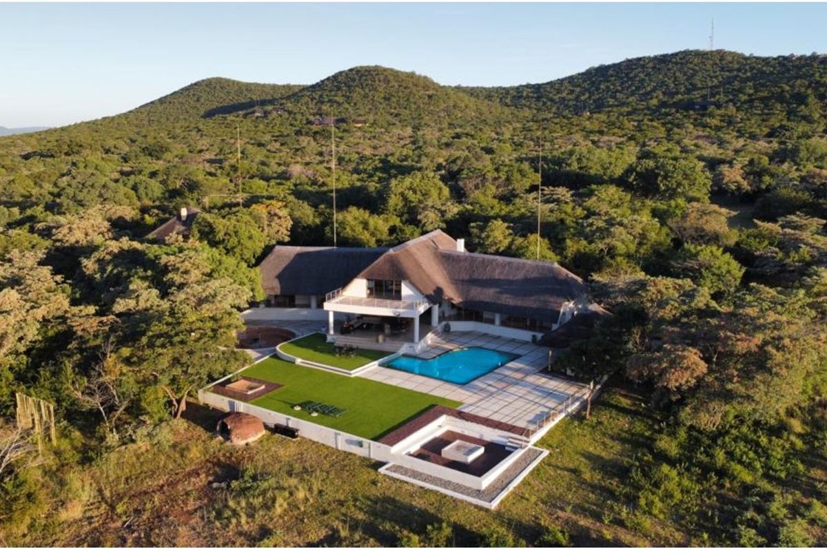 Aerial view of a large property featuring a thatched-roof house, swimming pool, expansive paved patio, and landscaped garden set against a natural bushveld backdrop.