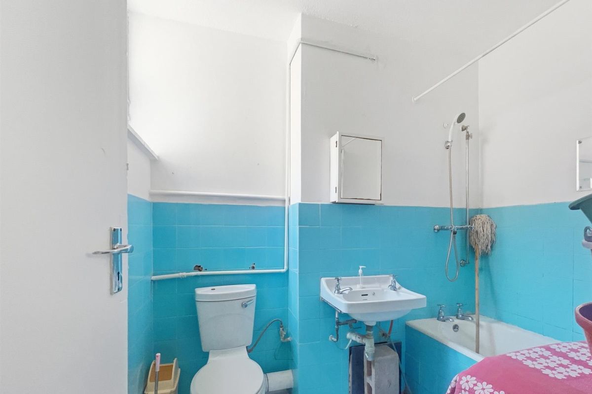 A dated bathroom featuring a standard toilet, wall-mounted basin with exposed plumbing, and a bathtub with a shower attachment.