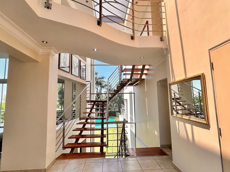 A bright, double-volume entrance hall featuring a modern floating timber staircase with stainless steel balustrades and large windows overlooking the garden and pool.