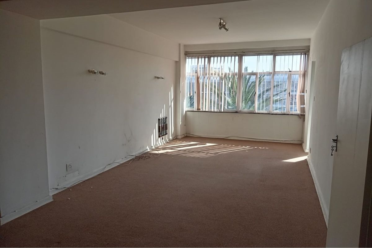 An empty, sunlit living room featuring carpet flooring, vertical blinds, and a wall-mounted heater, ready for renovation or staging.