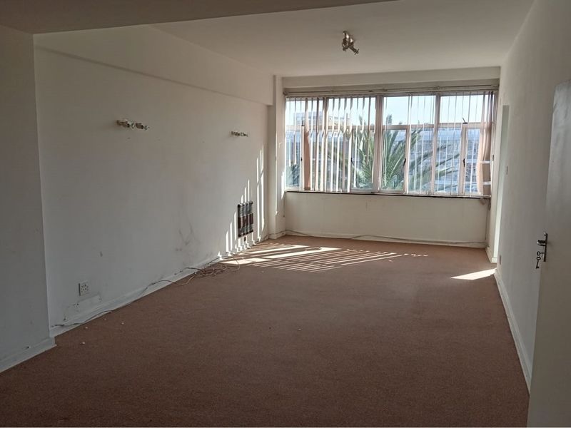 An empty, sunlit living room featuring carpet flooring, vertical blinds, and a wall-mounted heater, ready for renovation or staging.