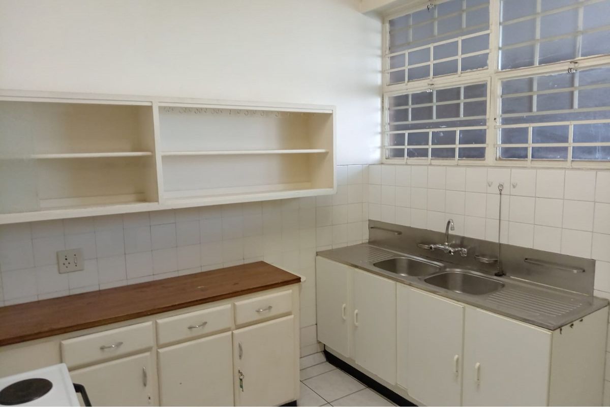A functional, empty kitchen featuring built-in cabinetry, a double bowl stainless steel sink, tiled splashbacks, and security bars on the window.