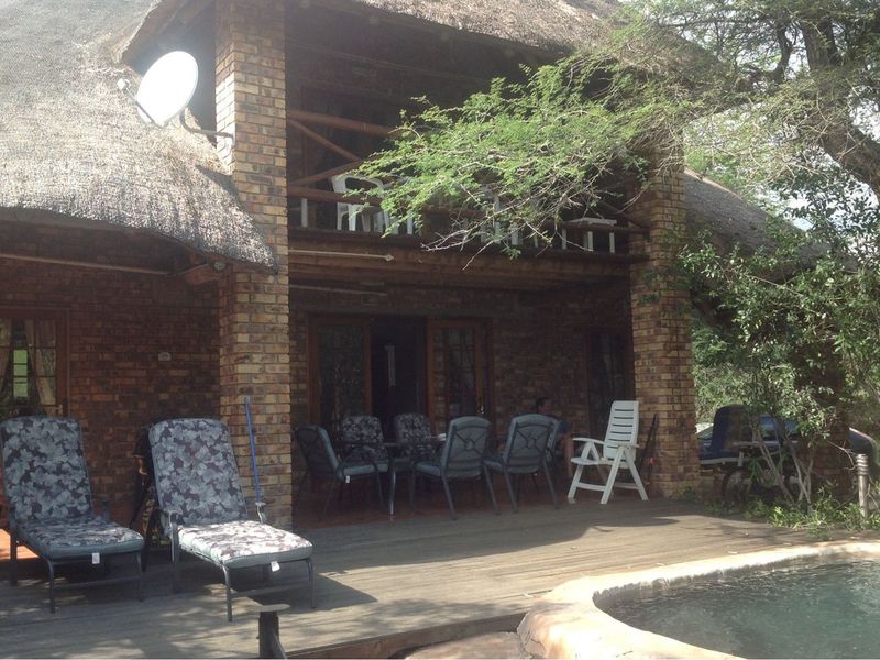 Rear exterior view of a rustic brick house with a thatched roof, featuring a wooden deck, swimming pool, and an upper-level balcony.