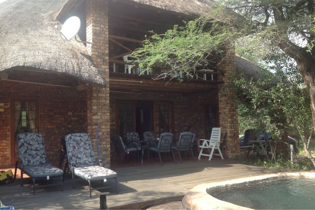 Rear exterior view of a rustic brick house with a thatched roof, featuring a wooden deck, swimming pool, and an upper-level balcony.