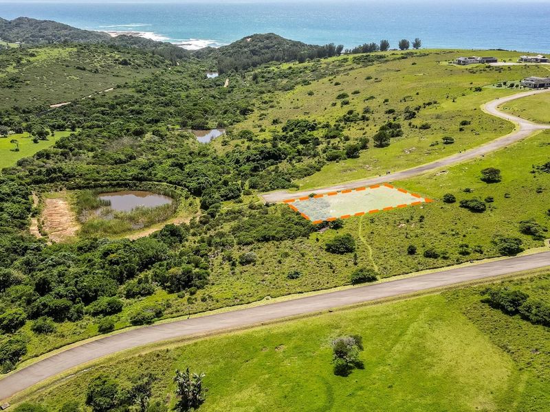 Aerial view of a vacant residential plot situated in a coastal estate, featuring paved road access and scenic natural surroundings.