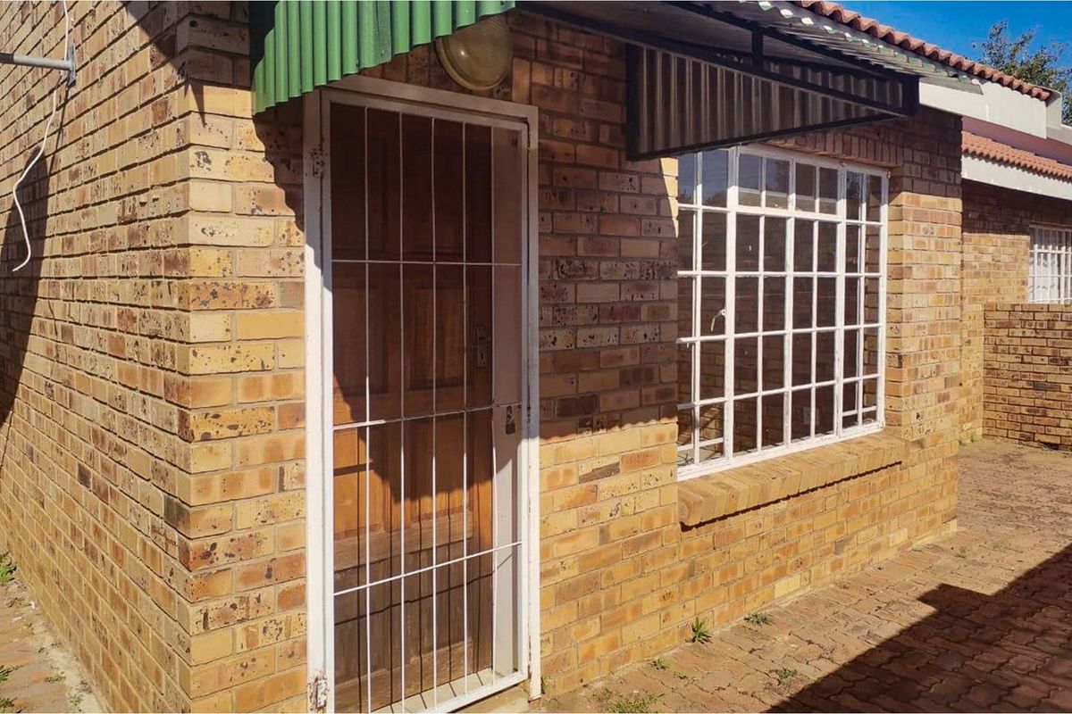 Exterior view of a face brick townhouse featuring a security gate, burglar bars, and a paved patio area.