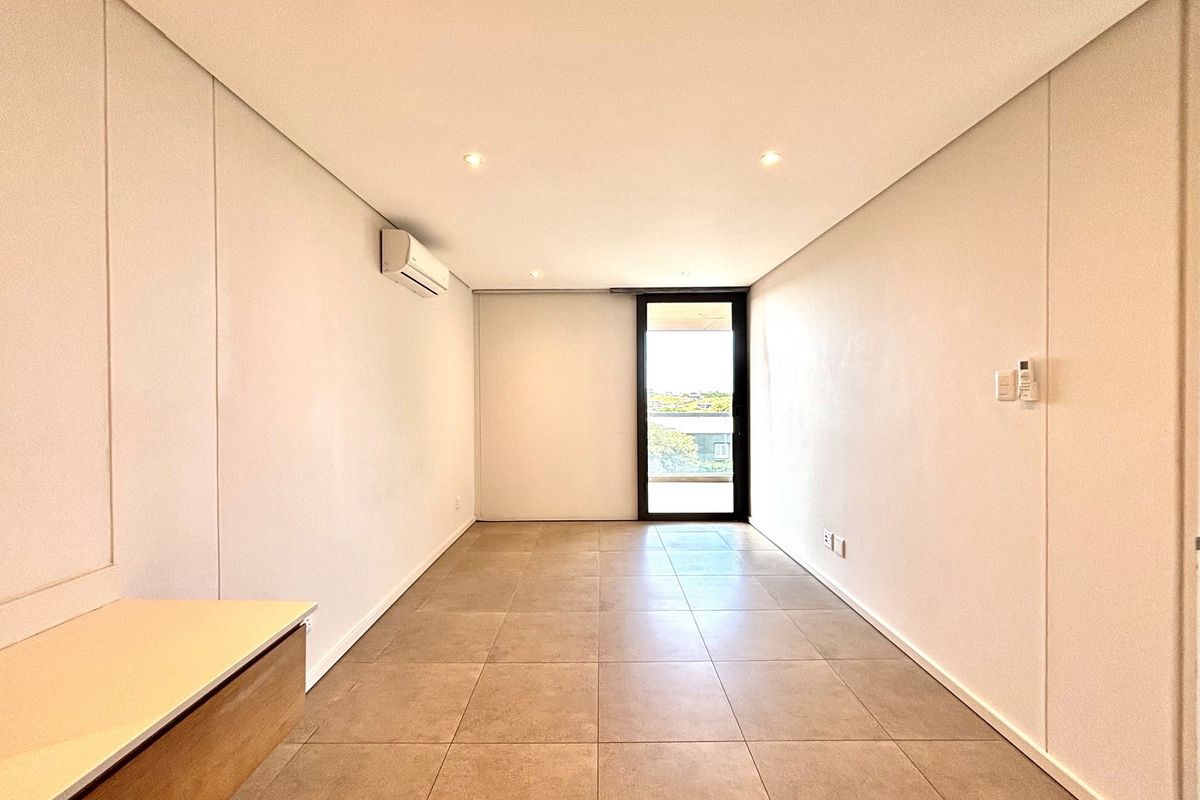 A bright, empty living area featuring tiled flooring, recessed ceiling lighting, a wall-mounted air conditioning unit, and a glass door leading to a balcony.