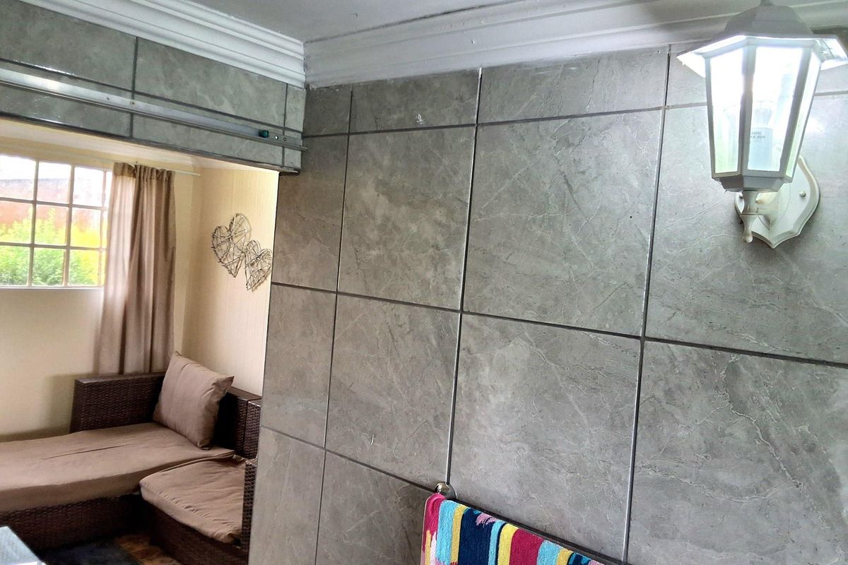 A living area featuring large format grey wall tiles, crown molding, and a wall-mounted light fixture, with an adjacent room visible through a sliding doorway.