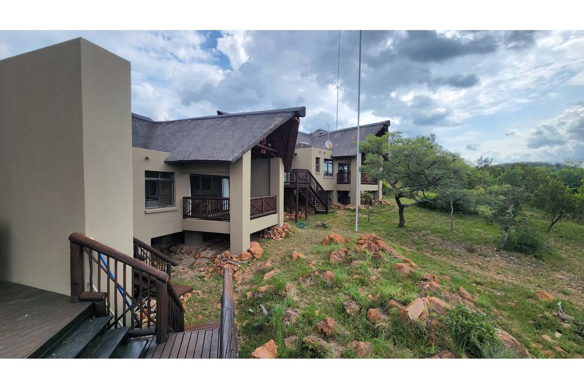 Rear view of a multi-level house featuring thatched roofing, wooden balconies, and stairs, set within a natural bushveld landscape.