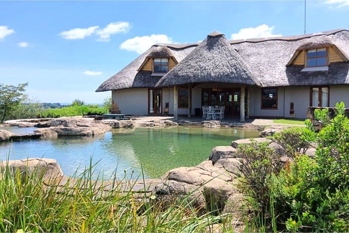 A scenic rear view of a thatched-roof house featuring a natural rock-lined swimming pool and a covered patio area.