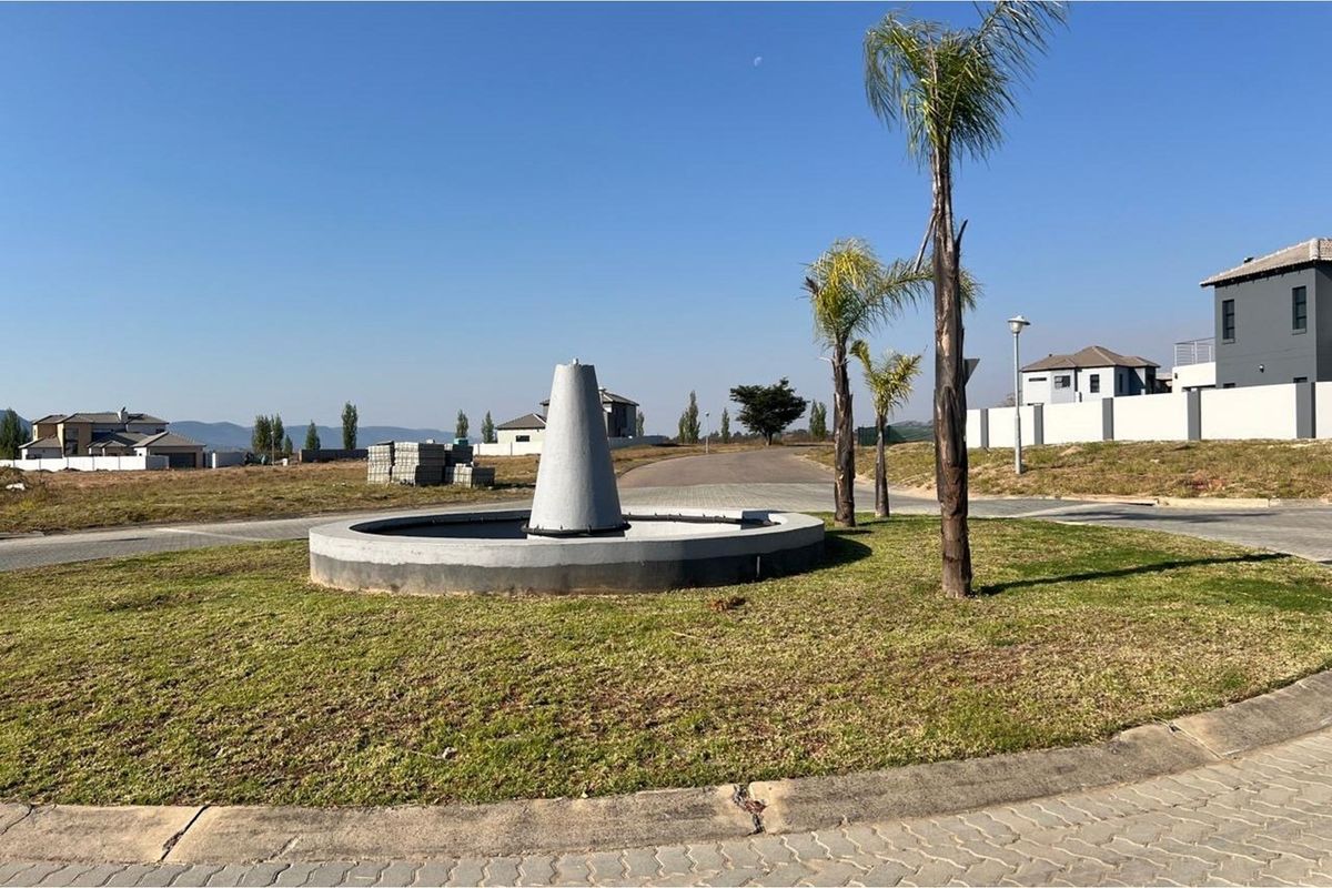 A view of a residential estate entrance featuring a central concrete fountain, paved roads, and landscaped garden areas with palm trees.