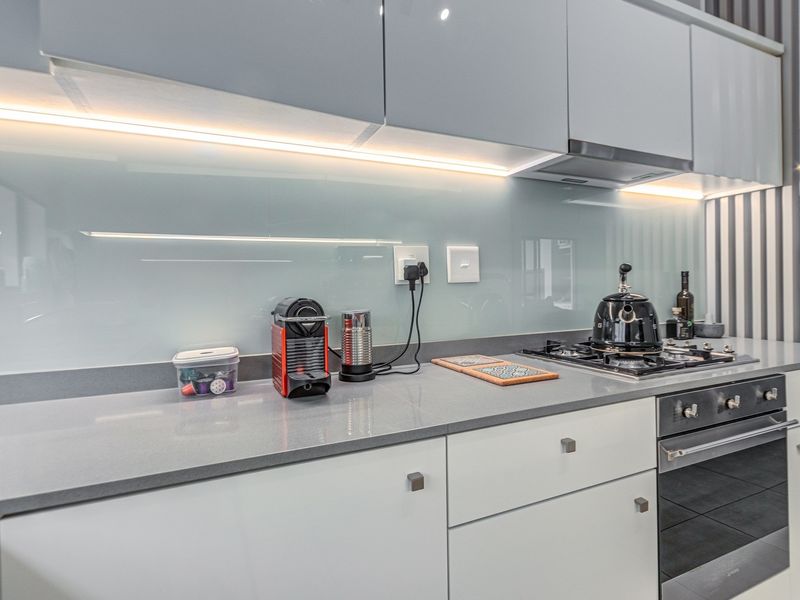 Modern kitchen featuring sleek white cabinetry, stone countertops, a glass splashback with integrated lighting, and high-quality built-in gas cooking appliances.