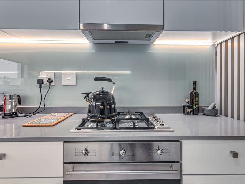Modern kitchen featuring a gas hob, built-in oven, extractor fan, sleek stone countertops, and a glass splashback with integrated under-cabinet lighting.