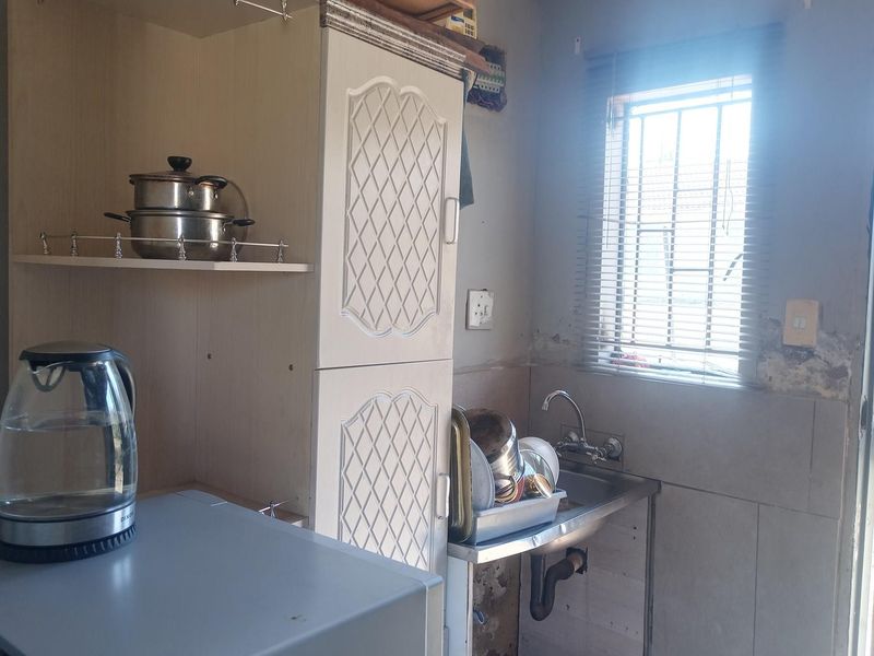 A functional scullery area featuring built-in cabinetry, a stainless steel sink, and a window with security bars, requiring some cosmetic wall repairs.