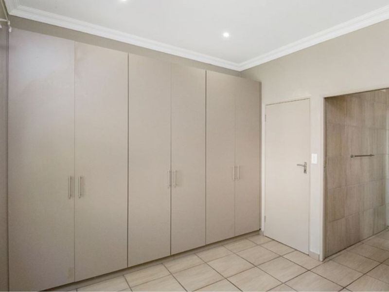 A clean, empty bedroom featuring extensive built-in cupboards, neutral tiled flooring, and recessed lighting, with an adjacent doorway leading to a tiled area.