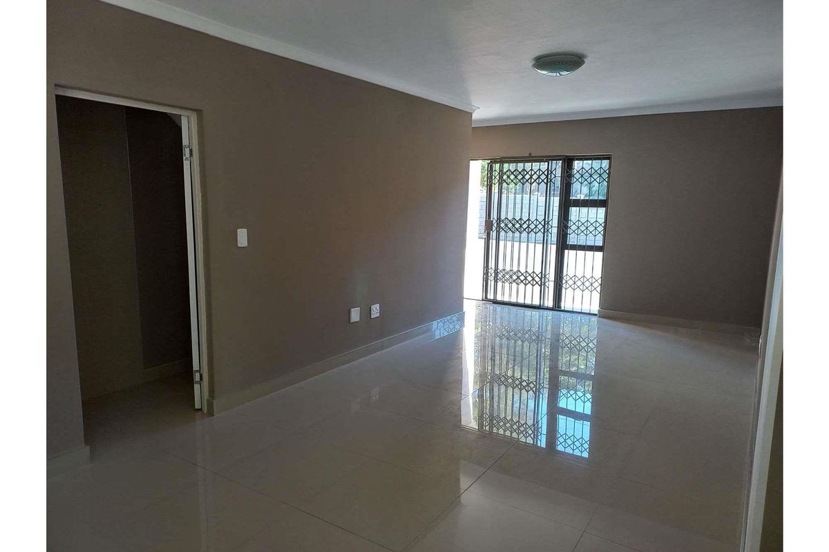 A spacious, empty living area featuring polished floor tiles, neutral wall tones, and a sliding glass door with security gate access to the exterior.