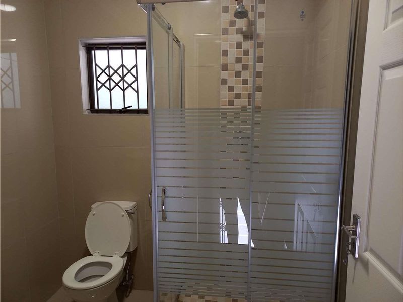 A functional bathroom featuring a glass-enclosed shower, tiled walls, and a toilet, with a window fitted with security bars.