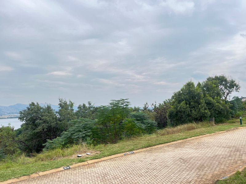 A view of a vacant residential plot featuring paved road access, established greenery, and scenic landscape views overlooking the water.