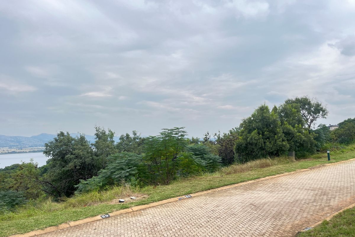 A view of a vacant residential plot featuring paved road access, established greenery, and scenic landscape views overlooking the water.