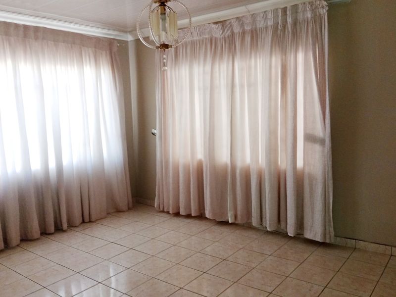A bright, empty living room featuring neutral tiled flooring, ceiling cornice detailing, and large windows dressed with curtains.