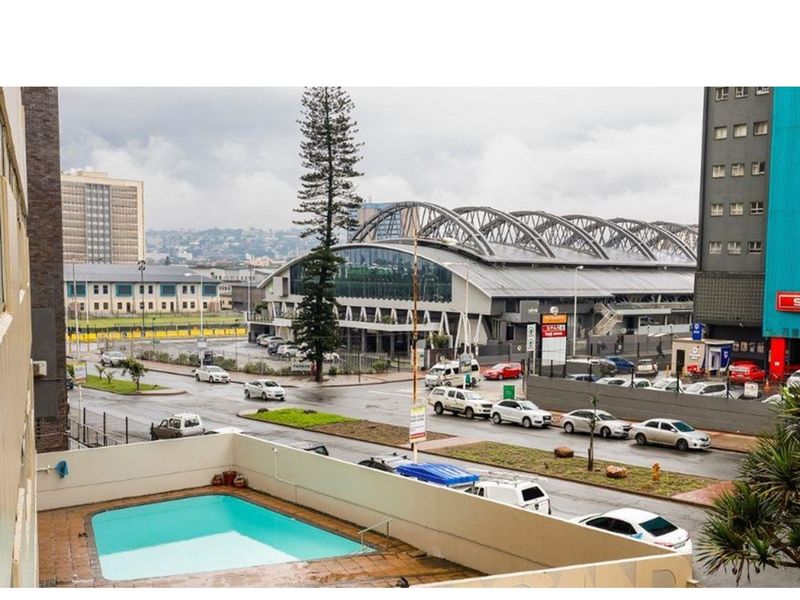 Elevated view overlooking a swimming pool with a surrounding terrace, featuring an urban cityscape and nearby commercial infrastructure in the background.